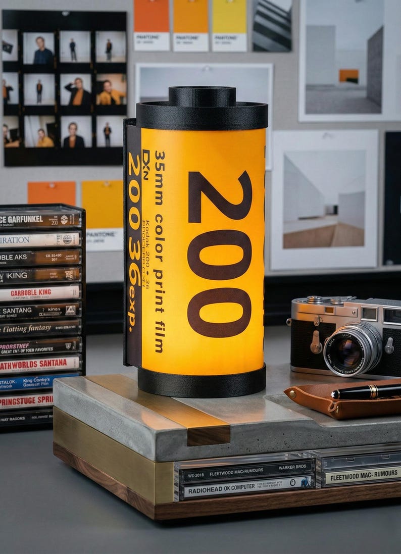 May include: A decorative lamp shaped like a 35mm Kodak film canister, with the number "200" prominently displayed. The lamp has a black top and base, and the film canister is yellow with black text. A vintage camera and cassette tapes are also visible.