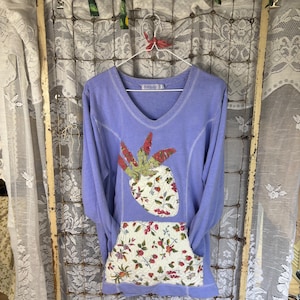 May include: A light purple long-sleeve shirt with a V-neckline, featuring a large quilted strawberry-shaped appliqué on the chest and a matching quilted front pocket, both decorated with a red and green floral and berry pattern.