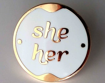 Könnte beinhalten: Eine weiße Emaille-Anstecknadel mit goldener Umrandung. Die Anstecknadel trägt die Aufschrift "she her" in goldenen Buchstaben.