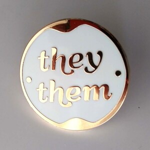 May include: A white enamel pin with a gold border and the words "they/them" in gold lettering.