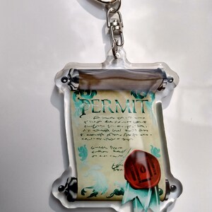 May include: A clear acrylic keychain featuring a scroll with the word "PERMIT" written on it. The scroll is decorated with a red wax seal and a green ribbon.
