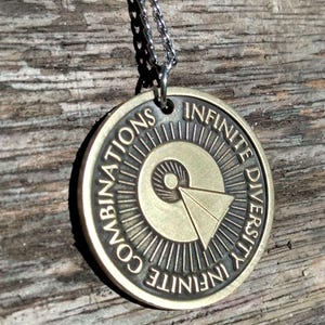 May include: A bronze-colored pendant necklace with the words "COMBINATIONS INFINITE DIVERSITY INFINITE" around the edge. The pendant has a circular design with a geometric shape in the center and is suspended from a silver chain.