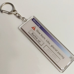 May include: A silver keychain with a blue and white warning message that reads "Warning: Slow burn. Like, glacial pacing. Buckle up. Just Kiss." The message is in a Windows error message style.