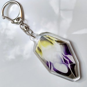 May include: A clear acrylic keychain with a purple and yellow geometric design. The keychain is shaped like a crystal and has a silver metal ring.