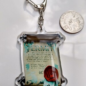 May include: A clear acrylic keychain featuring a scroll with the word "PERMIT" printed on it. The scroll is decorated with a red wax seal and a blue ribbon. The keychain is attached to a silver metal ring.