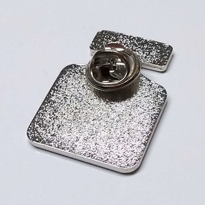 May include: Silver metal pin with a square shape and a pin back. The pin is blank.
