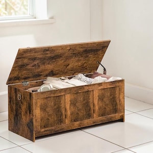 May include: A wooden storage chest with a hinged lid, open to reveal folded clothing and headphones. The chest has a distressed brown finish and is set against a white wall and floor.