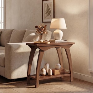 May include: A brown wooden console table with a curved apron and a lower shelf. The top surface holds a white table lamp with a fabric shade, a gold tray with dried flowers, and stacked books. The lower shelf holds three ceramic vases and a box.