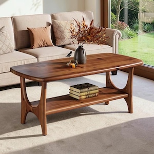 May include: A rectangular wooden coffee table with a smooth, polished finish and rounded edges. The table features a lower shelf holding three stacked books and a dark vase with dried foliage on the top surface, supported by four curved legs.