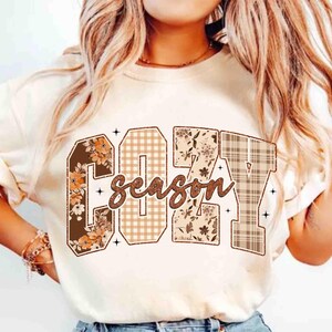 Peut inclure: T-shirt crème avec l'inscription "COZY SEASON" dans une police décorative. Les lettres sont remplies de motifs floraux, à carreaux et à motifs. Le t-shirt a des manches courtes.