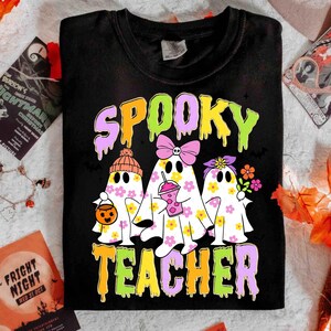 May include: Black t-shirt with the words "SPOOKY TEACHER" in dripping, colourful letters. The shirt features three ghost characters decorated with flowers, bows, and a pumpkin bucket, perfect for Halloween.