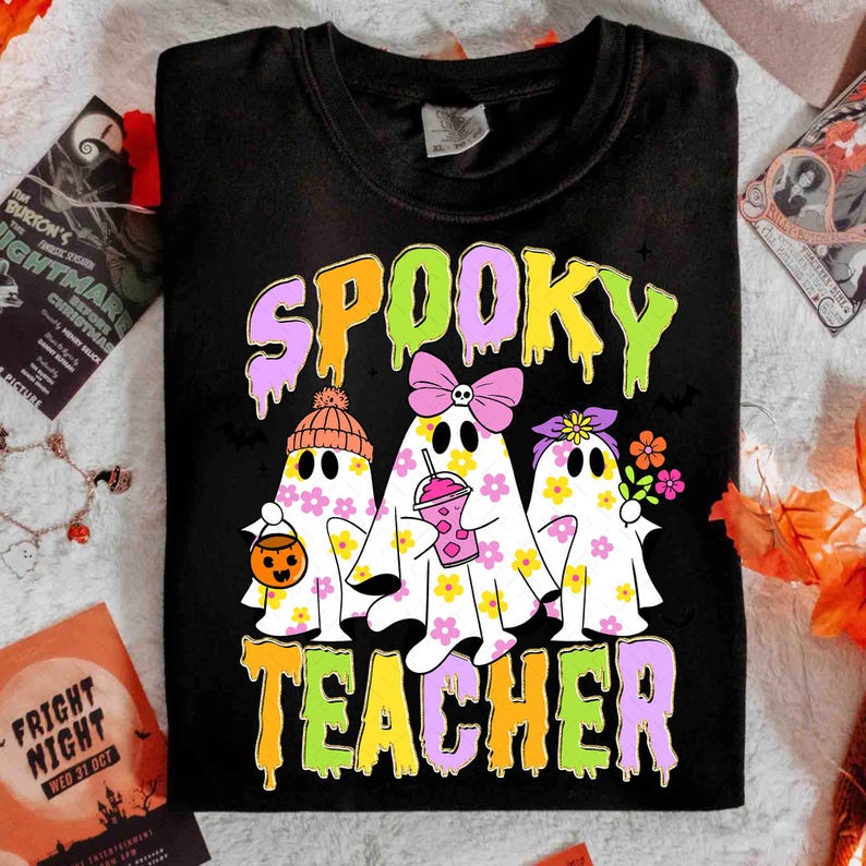 May include: Black t-shirt with the words "SPOOKY TEACHER" in dripping, colourful letters. The shirt features three ghost characters decorated with flowers, bows, and a pumpkin bucket, perfect for Halloween.