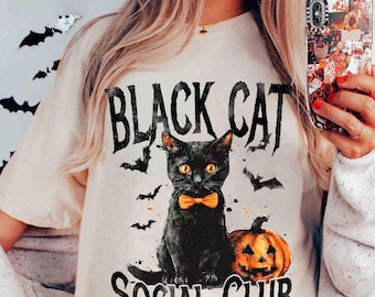May include: Cream-colored t-shirt featuring a black cat with an orange bow tie, sitting next to a carved pumpkin. The shirt has the text "BLACK CAT SOCIAL CLUB" and bat silhouettes. The cat has orange eyes.