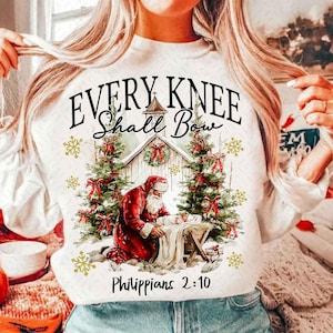 May include: White sweatshirt with a Christmas-themed graphic. The design features Santa Claus kneeling before baby Jesus in a manger, with text that reads "EVERY KNEE SHALL BOW" and "Philippians 2:10". The design includes Christmas trees and snowflakes.