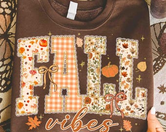 May include: A brown sweatshirt with the words "FALL VIBES" in a fall-themed design. The letters are decorated with pumpkins, leaves, and floral patterns. The letter "A" has an orange gingham pattern. A bow is on the letter "A" and "L".