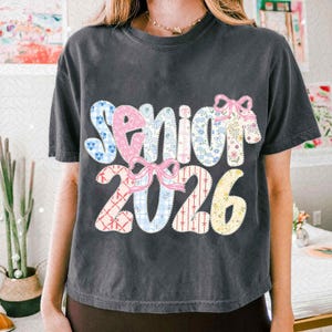 May include: Dark gray t-shirt with the word "Senior 2026" in a patchwork design. The text features various floral and patterned fabrics, with pink bows. A casual, comfortable tee.