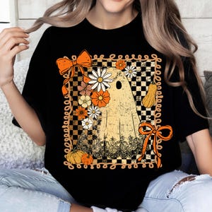 Puede incluir: Camiseta negra con un dise&ntilde;o de fantasma vintage adornado con flores, calabazas y lazos naranjas. El fantasma se encuentra sobre un fondo a cuadros con un borde decorativo, ideal para Halloween o el oto&ntilde;o.