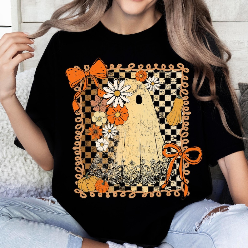 Puede incluir: Camiseta negra con un dise&ntilde;o de fantasma vintage adornado con flores, calabazas y lazos naranjas. El fantasma se encuentra sobre un fondo a cuadros con un borde decorativo, ideal para Halloween o el oto&ntilde;o.