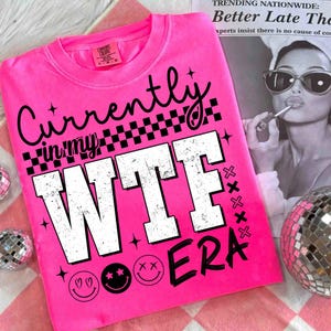 May include: A vibrant pink t-shirt featuring the phrase "Currently in my WTF ERA" in a distressed white font. The design incorporates black accents, smiley faces, stars, and a checkered pattern. The shirt is placed on a pink and white checkered surface.
