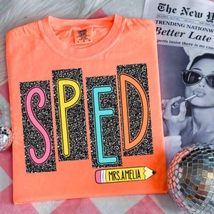 May include: Coral-colored t-shirt with the word "SPED" in colorful, glitter-filled letters. A pencil graphic with the text "MRS.AMELIA" is below the letters. The shirt is on a pink and white checkered surface, with a newspaper and disco ball in the background.