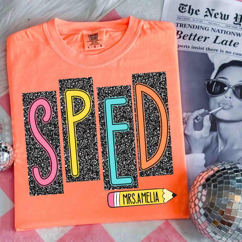 May include: Coral-colored t-shirt with the word "SPED" in colorful, glitter-filled letters. A pencil graphic with the text "MRS.AMELIA" is below the letters. The shirt is on a pink and white checkered surface, with a newspaper and disco ball in the background.