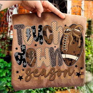 May include: A brown t-shirt featuring the words "Touch Down Season" in a mix of patterned fonts and a football graphic. The design includes leopard print, stars, and lightning bolts. The football has a brown bow.