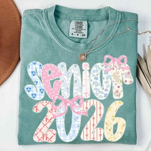 May include: A teal Comfort Colors t-shirt with the words "Senior 2026" in a patchwork design. The letters and numbers are decorated with floral and patterned fabrics, and pink bows. A gold necklace with a pendant is draped across the shirt.