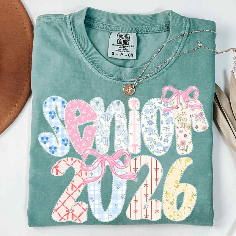 May include: A teal Comfort Colors t-shirt with the words "Senior 2026" in a patchwork design. The letters and numbers are decorated with floral and patterned fabrics, and pink bows. A gold necklace with a pendant is draped across the shirt.