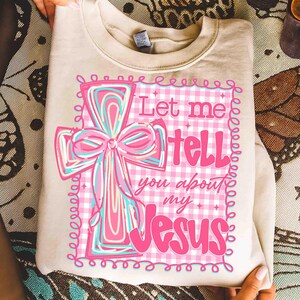 May include: Cream-colored sweatshirt featuring a pink and blue cross design with a bow. The cross is set against a pink and white gingham background, with the text "Let me tell you about my Jesus" in pink.