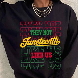 May include: Black sweatshirt featuring the words "THEY NOT" in red, "THEY NOT" in green, and "Juneteenth" in yellow. The text "LIKE US" is in green and red. A gold necklace is visible.