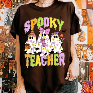 May include: A brown t-shirt with the words "Spooky Teacher" in a colourful, layered font. The shirt features three ghost characters decorated with flowers, bows, and accessories, perfect for Halloween.