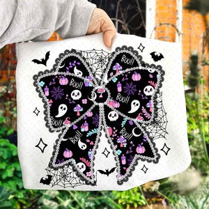 May include: A white Halloween-themed bow-shaped design with black, purple, and pink elements. The bow features skulls, ghosts, pumpkins, and the word "Boo!!" in a decorative font. The design is framed by a black lace-like border and spiderwebs.