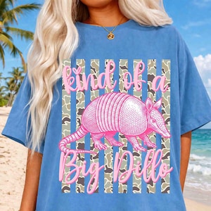May include: A light blue t-shirt with a pink armadillo graphic and the text "kind of a Big Dillo". The design features a camouflage pattern with vertical stripes. The shirt is made of a soft, comfortable material.