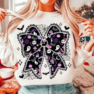 May include: White sweatshirt featuring a large black bow design with Halloween-themed illustrations. The bow includes skulls, pumpkins, mushrooms, and the word "Boo!!" in a playful font. The design is surrounded by a spiderweb and bats.