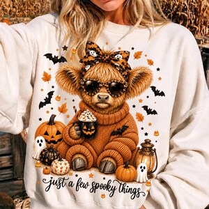 May include: A cream-coloured sweatshirt featuring a graphic of a fluffy Highland cow wearing a leopard print bow and sunglasses, holding a jack-o'-lantern mug. The design includes pumpkins, ghosts, bats, and the text "just a few spooky things."