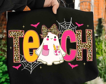 Może przedstawiać: Czarna koszulka z napisem "TEACH" w panterkę, żółtym i różowym. Postać ducha z kokardą w panterkę, trzymająca ołówki i nosząca różową kieszeń znajduje się na środku. Różowe nietoperze i pajęczyny dopełniają projekt.