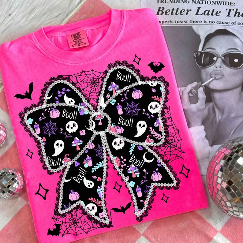 May include: A bright pink t-shirt featuring a large black bow design with a spooky Halloween theme. The bow is decorated with skulls, ghosts, pumpkins, and the word "Boo!!". The shirt has a crew neck and short sleeves, perfect for the Halloween season.