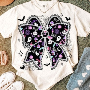 May include: Cream-colored t-shirt featuring a large black bow design with Halloween-themed illustrations. The bow includes skulls, ghosts, pumpkins, and the word "Boo!!". The design is outlined with a black lace trim and surrounded by spiderwebs and bats.