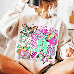 Könnte beinhalten: Ein weißer Pullover mit einem bunten Grafikdesign, auf dem "T-ball Mama" mit Baseballs, Schlägern und Cheerleader-Pompons steht.