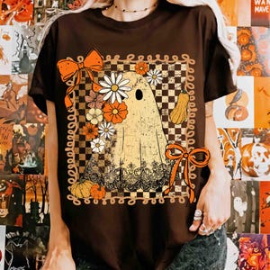 Puede incluir: Camiseta marr&oacute;n con un dise&ntilde;o de fantasma retro adornado con flores, calabazas y lazos. El fantasma tiene una corona floral y est&aacute; sobre un fondo a cuadros. El dise&ntilde;o utiliza tonos naranja, blanco y amarillo.