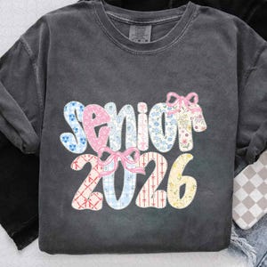 May include: Dark gray t-shirt with the word "Senior 2026" in a patchwork design. The word "Senior" is in pink, blue, and floral patterns, with pink bows. The numbers "2026" are in patchwork patterns of red, blue, and yellow.