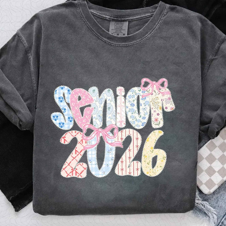 May include: Dark gray t-shirt with the word "Senior 2026" in a patchwork design. The word "Senior" is in pink, blue, and floral patterns, with pink bows. The numbers "2026" are in patchwork patterns of red, blue, and yellow.