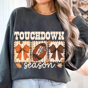 May include: Dark gray sweatshirt with the words "TOUCHDOWN season" in white script. The design includes a football, bows, and fall leaves in shades of orange and brown. The sweatshirt is a comfortable, casual style.