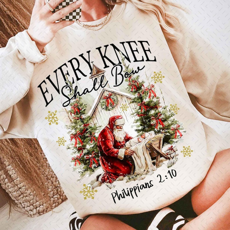 May include: Cream-colored sweatshirt with a Christmas-themed graphic. The design features Santa Claus kneeling, a nativity scene, Christmas trees, and the text "EVERY KNEE Shall Bow Philippians 2:10". Gold snowflakes are scattered around the image.