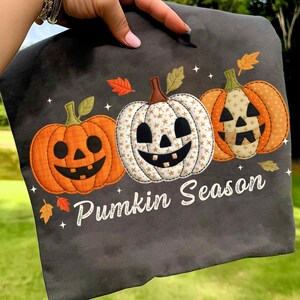 Peut inclure: T-shirt gris foncé avec trois citrouilles souriantes. Les citrouilles sont orange et blanches, avec des feuilles et le texte "Punkin Season". Un design festif pour l'automne.