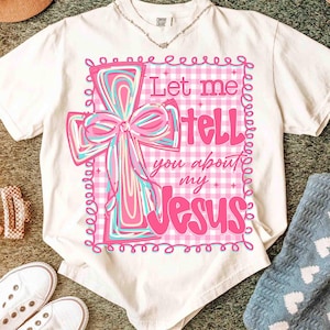 May include: Cream-colored t-shirt featuring a pink cross design with a bow and the text "Let me tell you about my Jesus." The cross is set against a pink and white gingham pattern, with a decorative border.