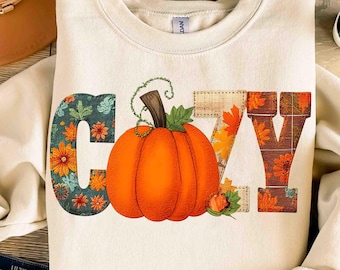 Puede incluir: Sudadera color crema con la palabra "COZY" en un diseño de otoño. La letra "C" es floral, la "O" es una calabaza naranja, la "Z" tiene hojas de otoño y la "Y" es floral. Una sudadera con temática otoñal.
