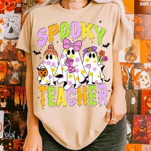 May include: A beige t-shirt with the words "Spooky Teacher" in a colourful, dripping font. The shirt features three ghost characters decorated with flowers, bows, and accessories, ideal for Halloween.