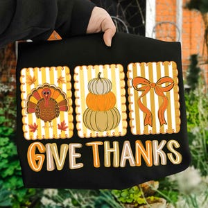 May include: Black Thanksgiving-themed tote bag with three rectangular panels. The panels feature a turkey, stacked pumpkins, and a bow, all set against a yellow and white striped background. The words "GIVE THANKS" are printed in orange and beige.