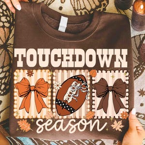 May include: Brown sweatshirt with the words "TOUCHDOWN season" in white. The design includes a football, bows, and fall leaves in orange, brown, and beige. The sweatshirt is held by hands.